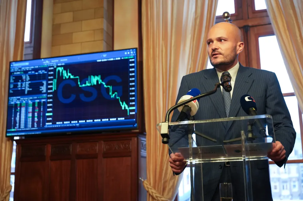 A businessman speaking at a podium with microphones, next to a screen showing financial market data.