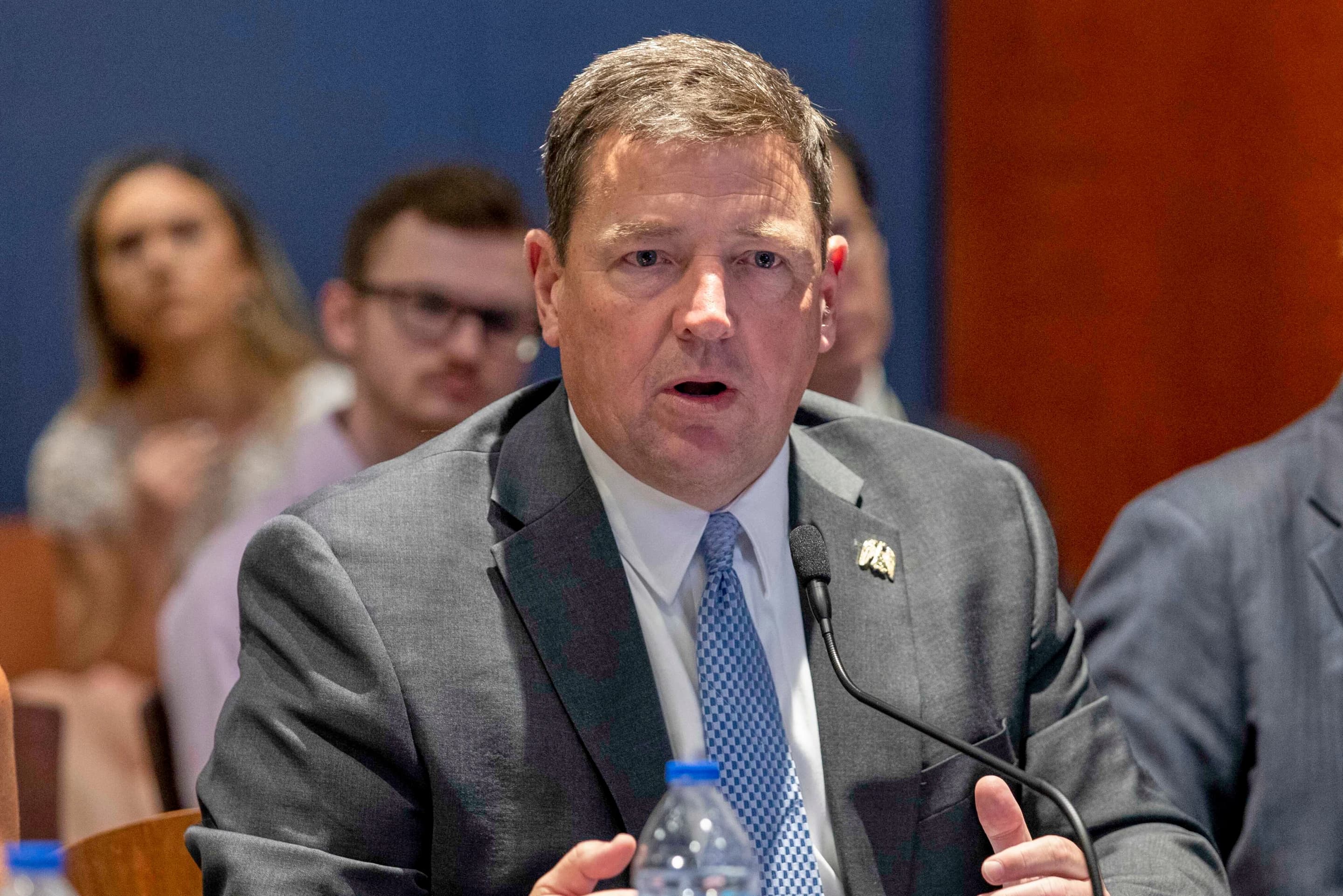 A Justice Department official speaking into a microphone during a hearing, with a blue and red backdrop behind him.