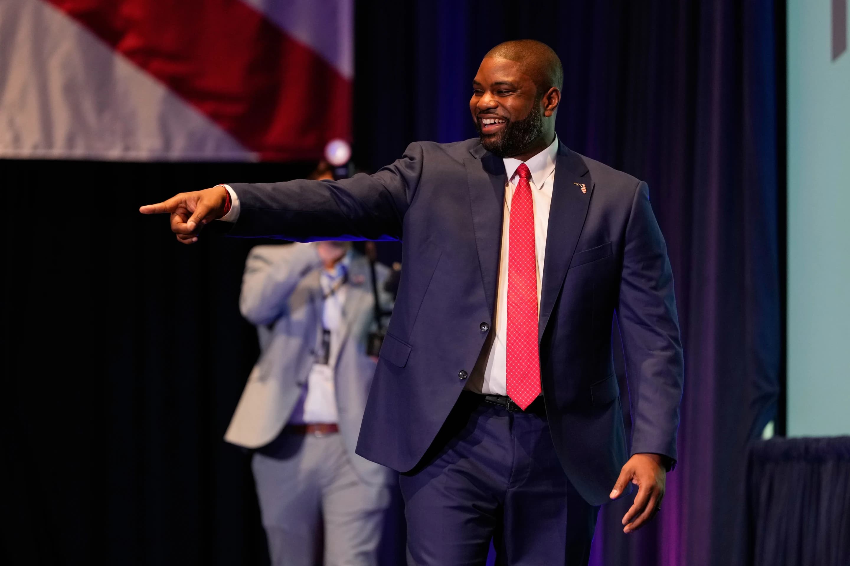 A Republican candidate pointing and smiling on stage at a state party event in Florida, with red white and blue bunting in the background.