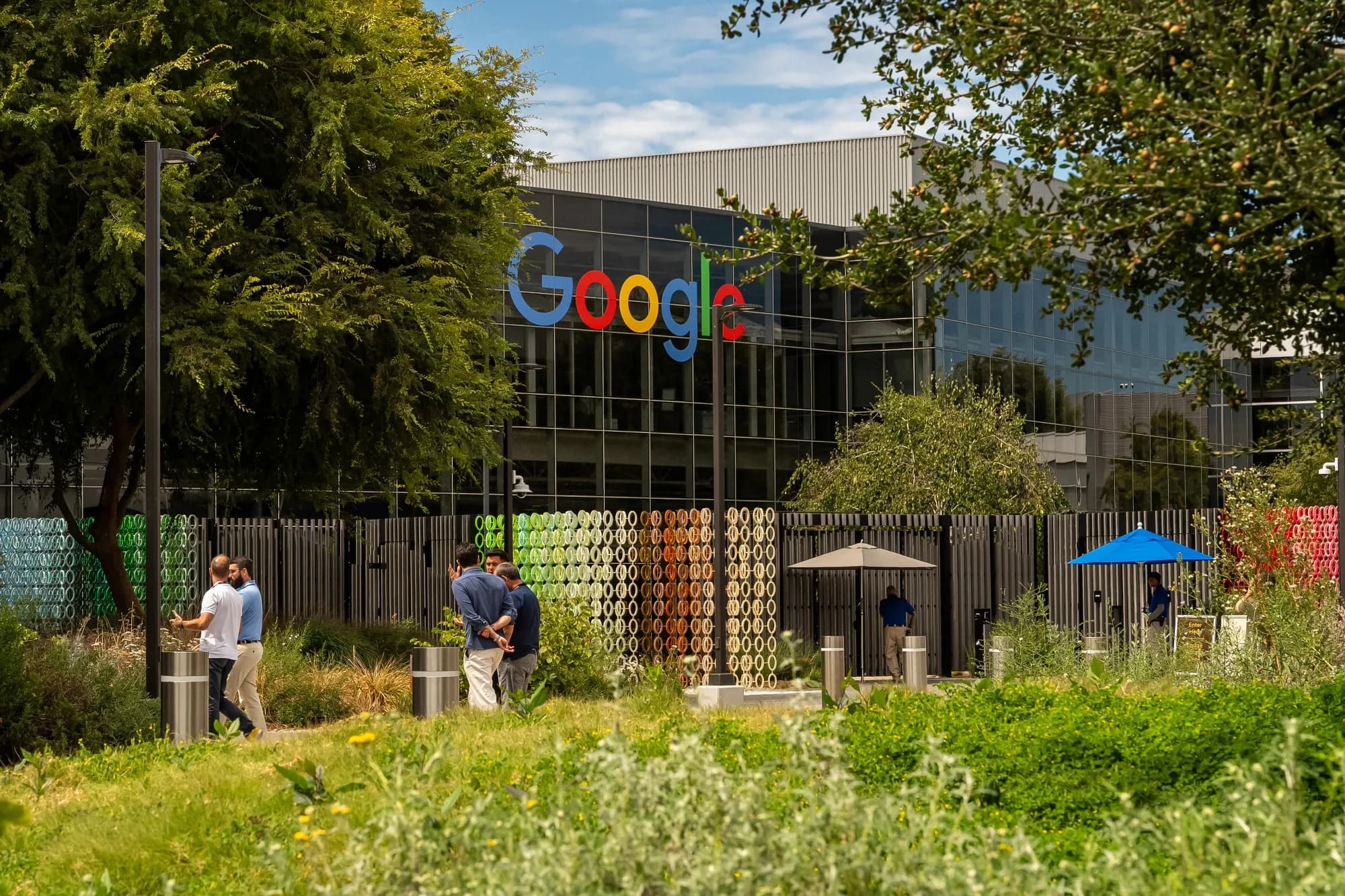 The Google logo displayed on the exterior of a corporate campus building, with people walking through a landscaped courtyard.