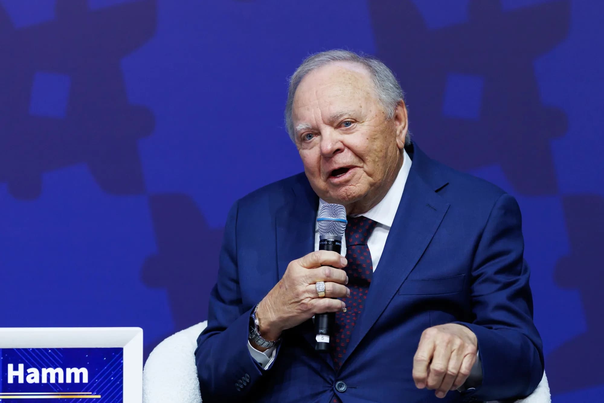 Harold Hamm speaking into a microphone during a panel discussion, seated in front of a blue backdrop.
