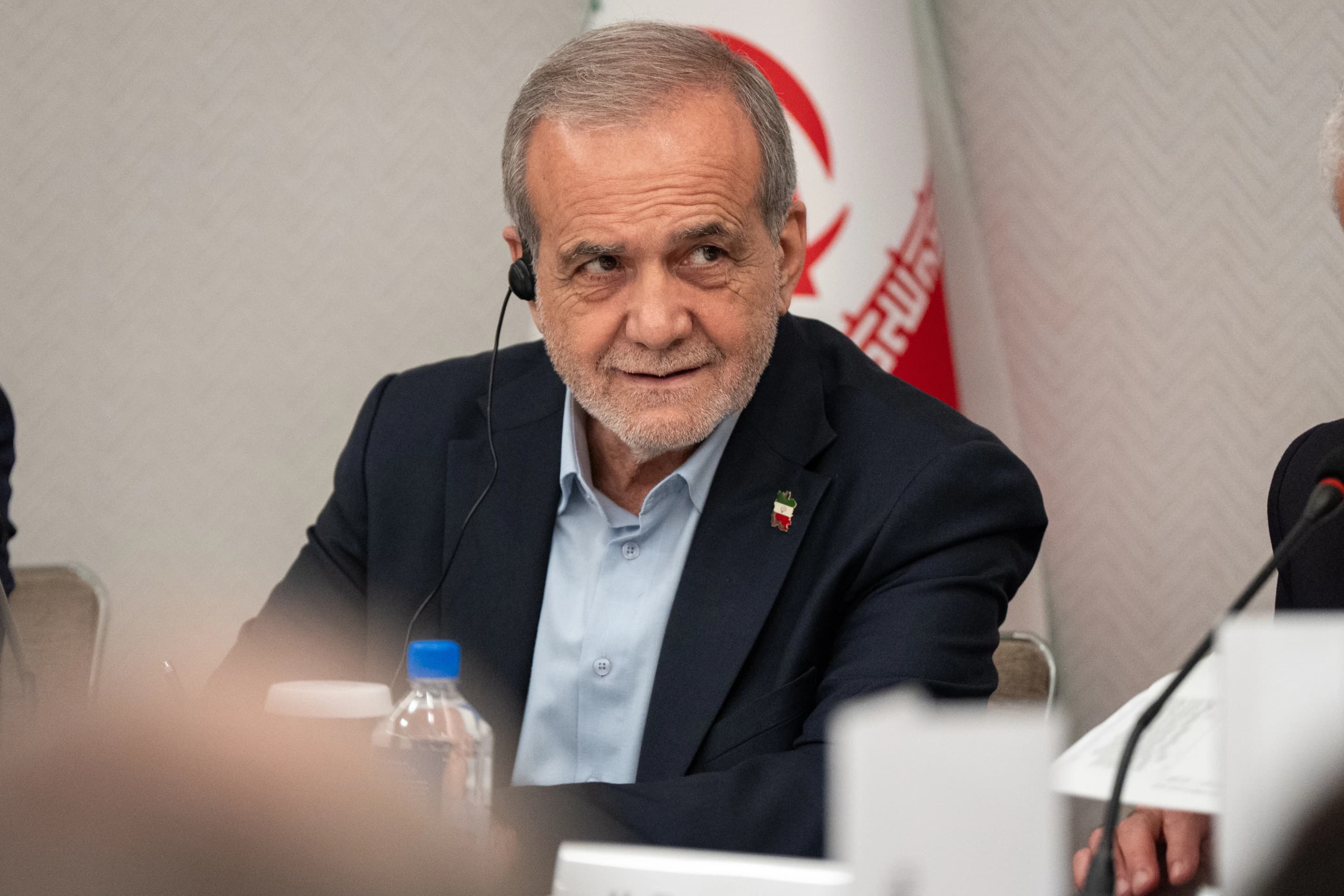 Iranian President Masoud Pezeshkian wearing an earpiece while seated at a conference table, with an Iran flag banner behind him.