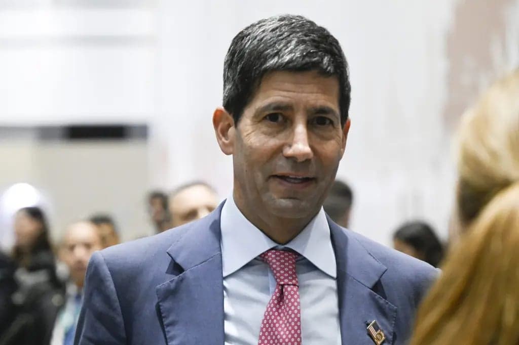 A man in a blue suit and pink tie speaking among a group of people indoors