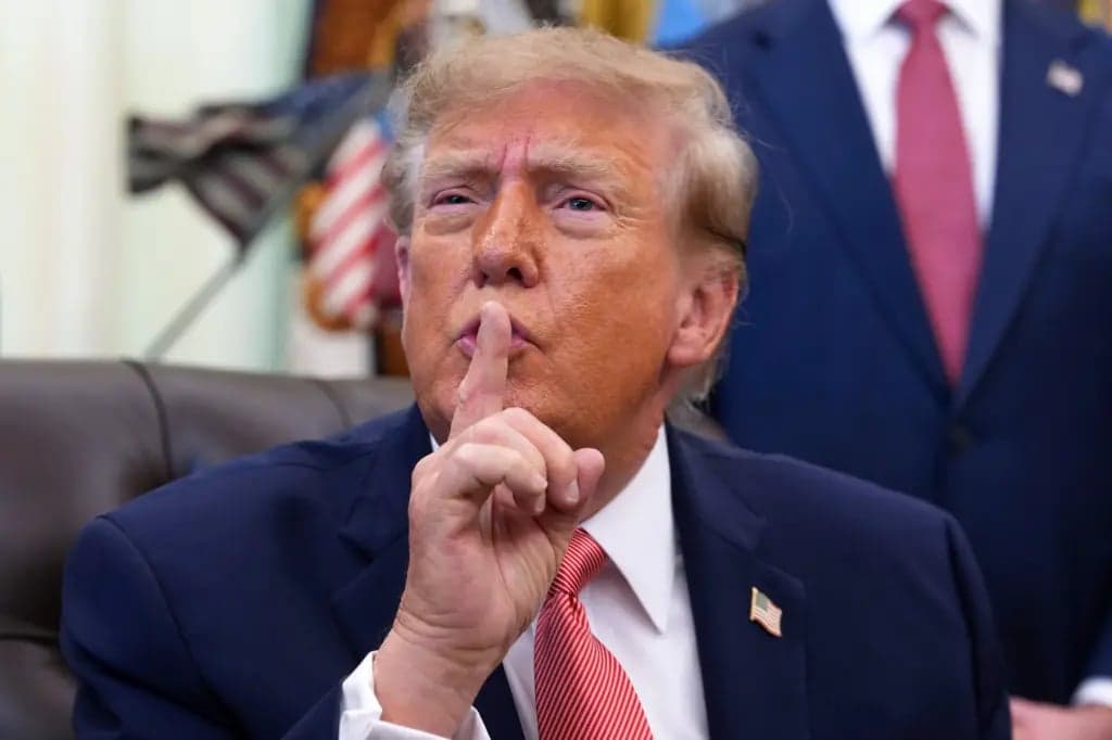 President Trump holding a finger to his lips in a shushing gesture while seated in the Oval Office