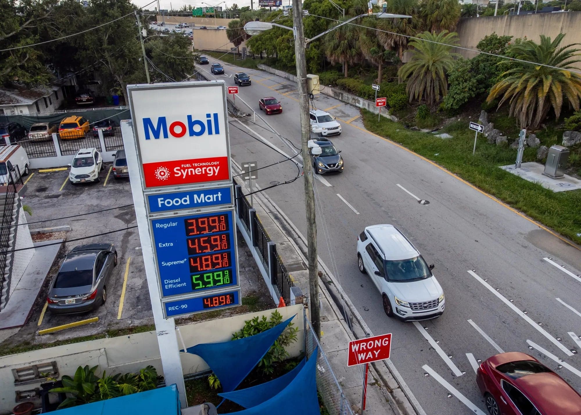 A Mobil gas station price sign showing regular, extra, supreme, and diesel prices, with cars driving past on the street below