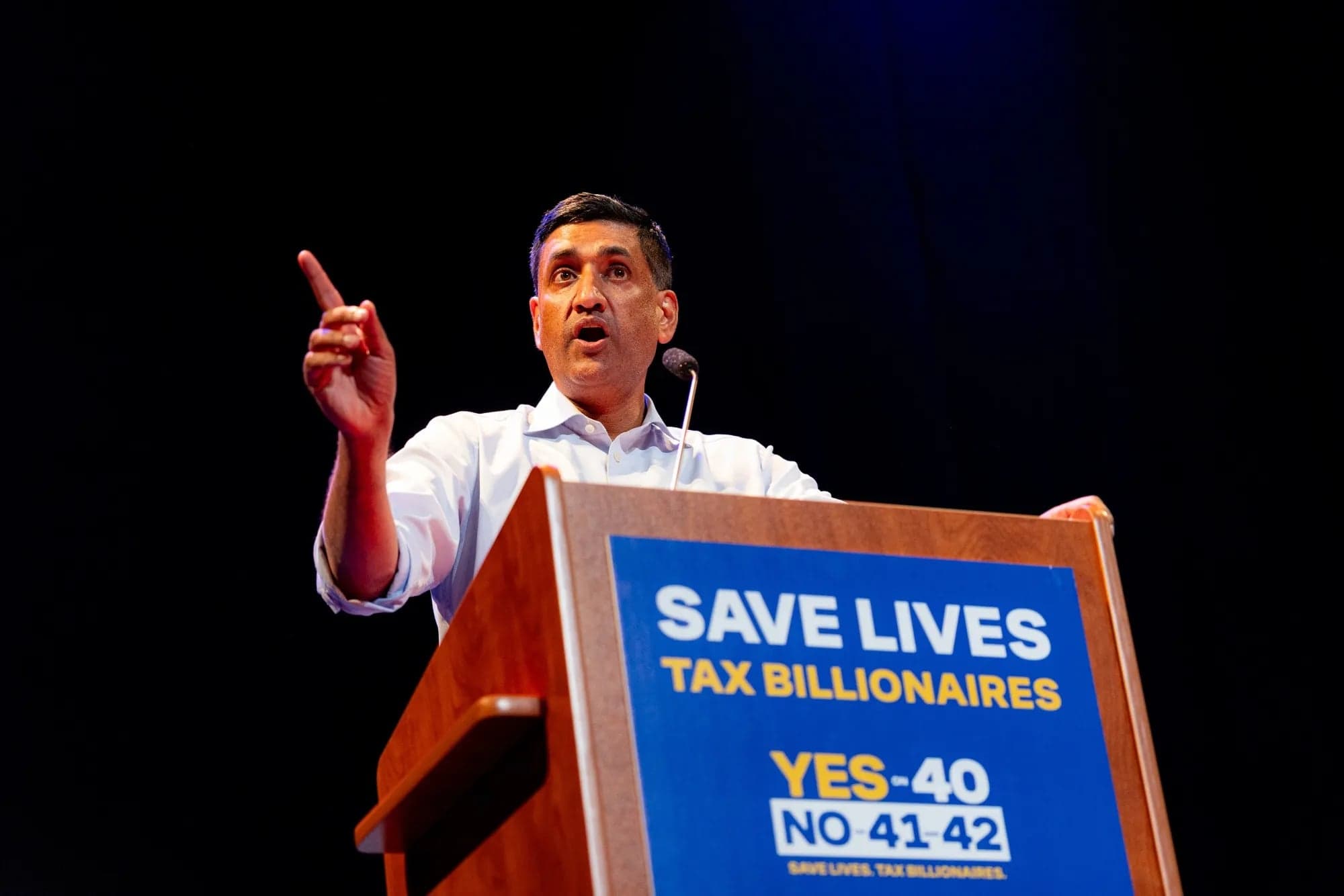 Rep. Ro Khanna speaking and pointing while standing at a podium with Save Lives Tax Billionaires signage