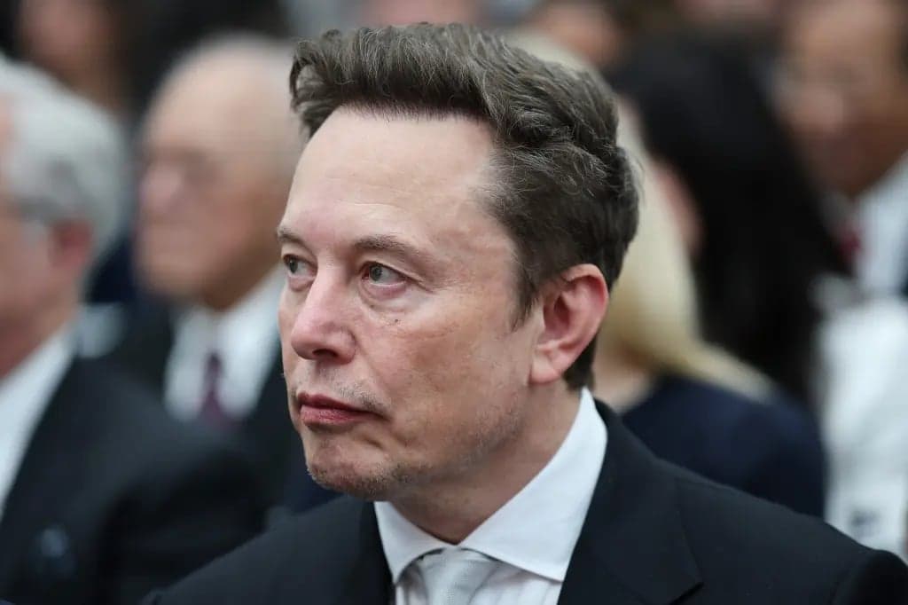 Elon Musk looking off to the side in a crowd, wearing a dark suit and light tie