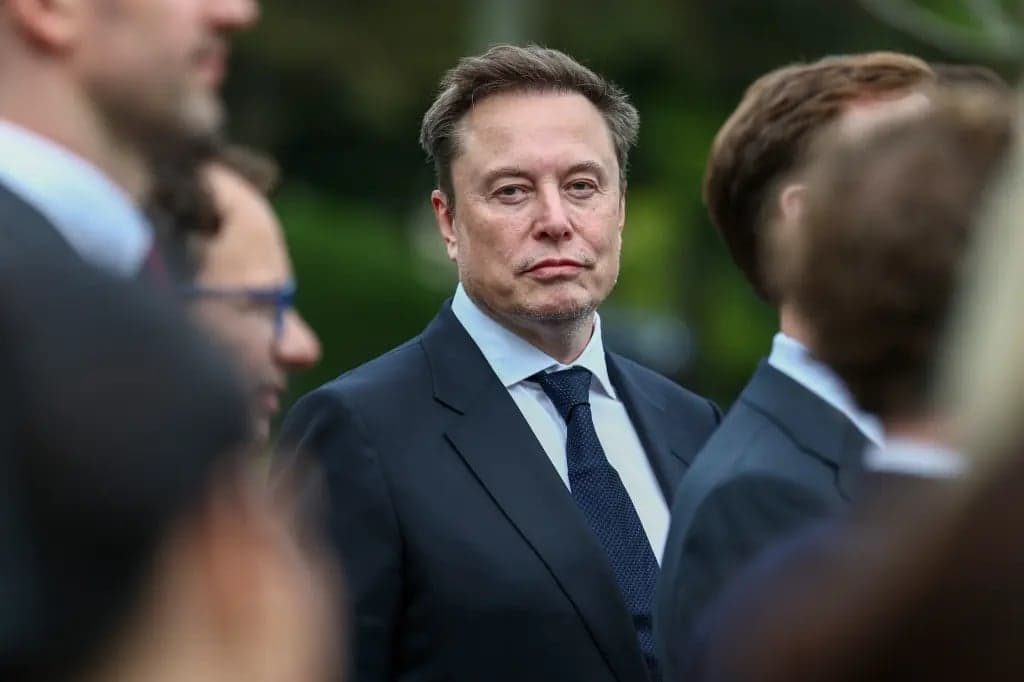 Elon Musk standing among a group of people outdoors, wearing a dark suit and tie